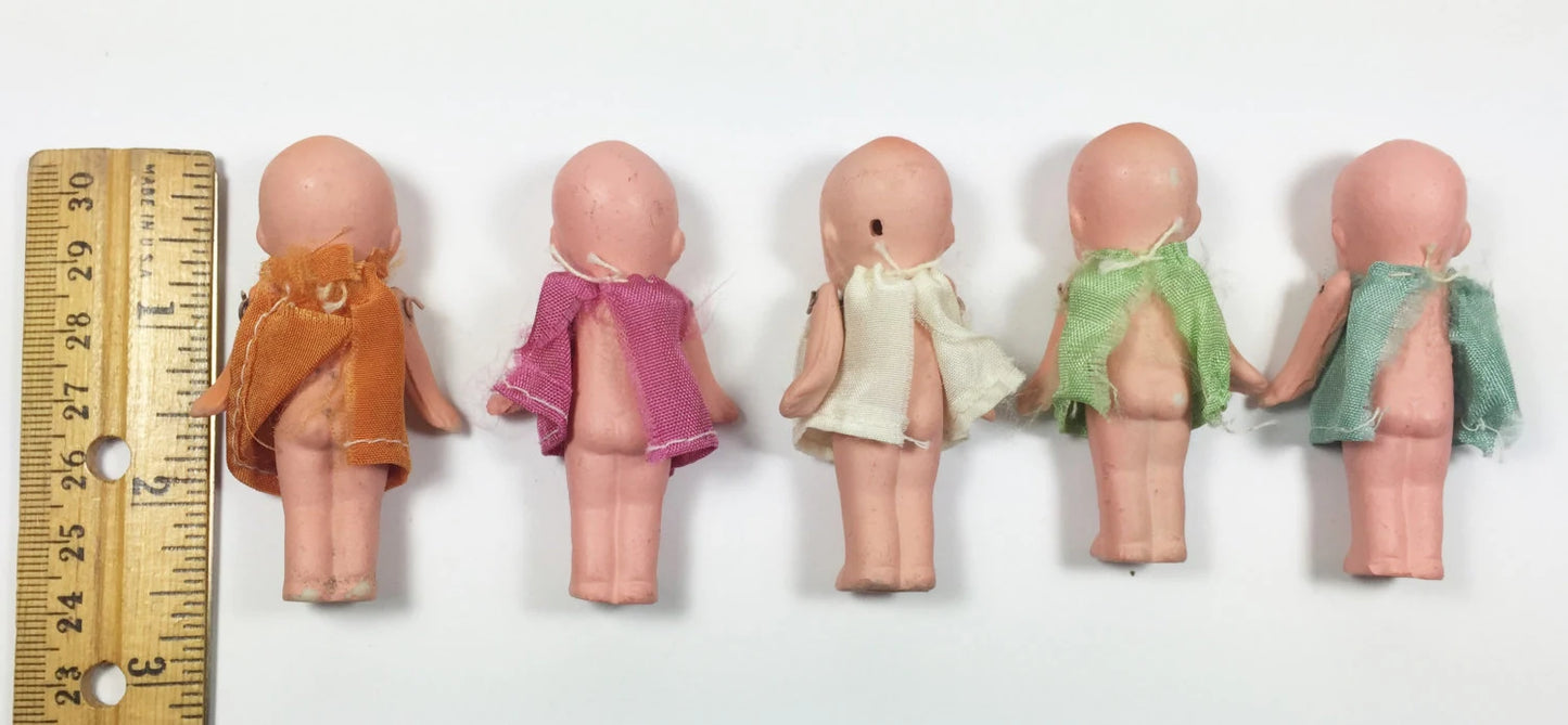 Five small dolls in various colored dresses lined up next to a ruler on a white background