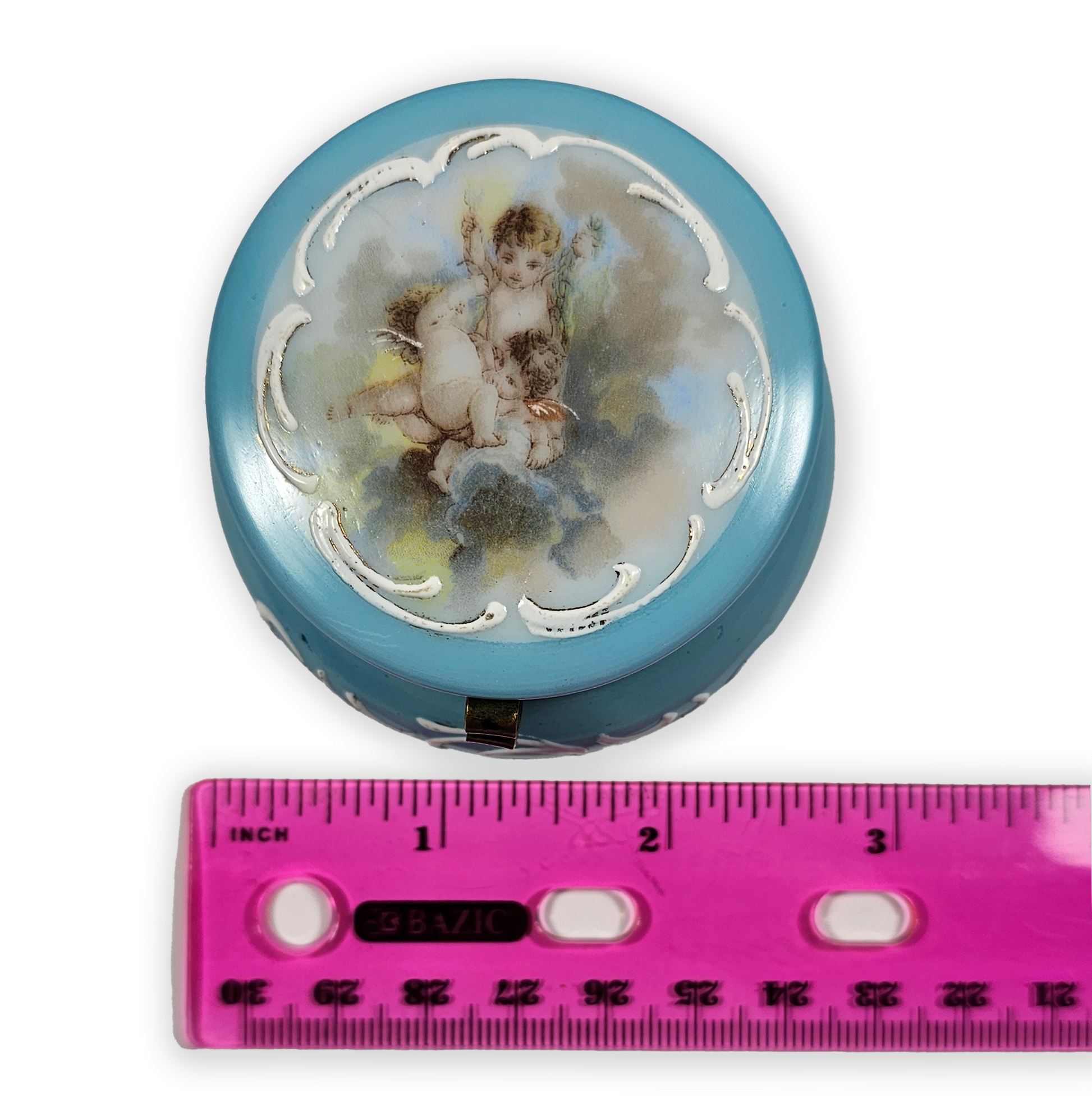 Round decorative item with cherub design next to a pink ruler for scale
