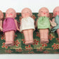Five small dolls with different colored dresses on a floral-patterned box.